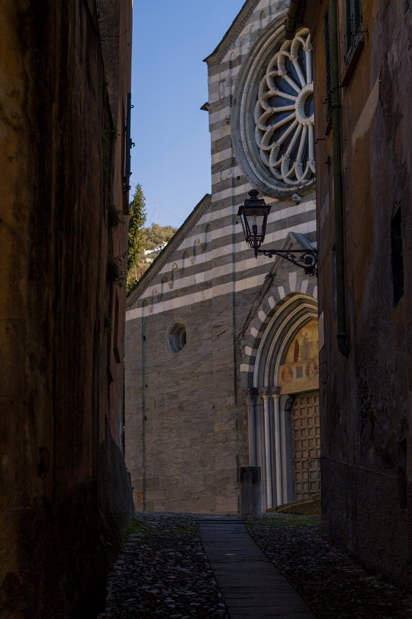 A narrow alleyway with stone buildings, a large arched doorway, and a circular window on the right.