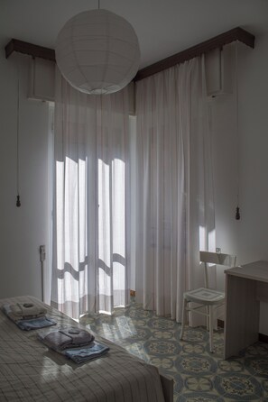 A bedroom with a bed, a desk, and a chair. There is a window with sheer curtains and a hanging light fixture.