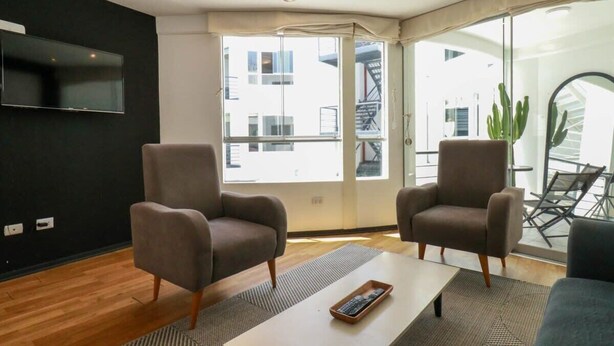 Una sala de estar moderna con dos sillones grises, una mesa de centro blanca y un televisor de pantalla plana montado en una pared negra.