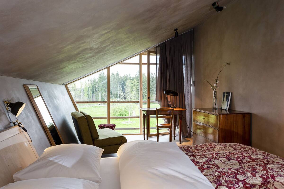 Une chambre avec une grande fenêtre, un bureau, une chaise, un lit et une table de chevet.