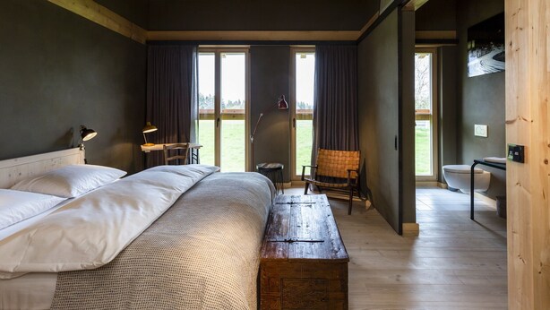 A bedroom with a large bed, a wooden bench, a desk, and a chair. There are two windows with curtains, a door leading to a bathroom, and a view of a grassy area outside. - Gut Sonnenhausen (Glonn)