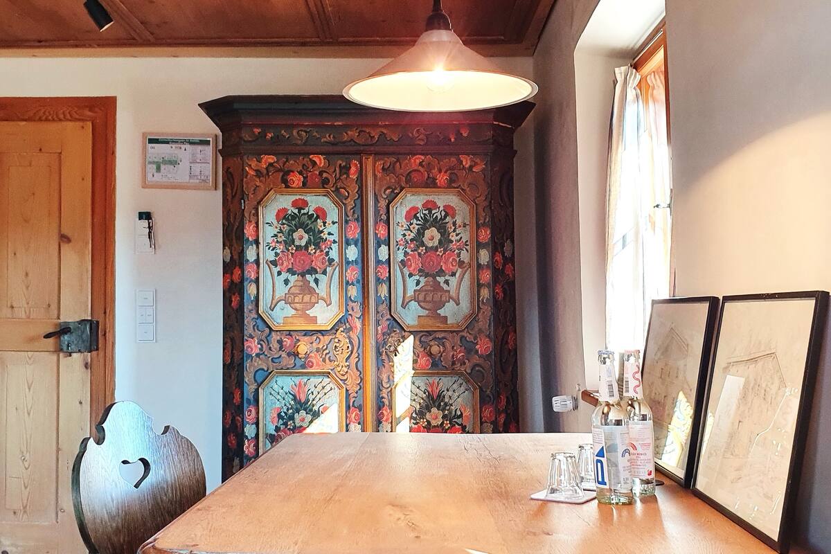 Une table de salle à manger en bois, ornée d’une bougie, d’une bouteille et de verres. Derrière la table, on aperçoit une porte décorative aux motifs floraux.