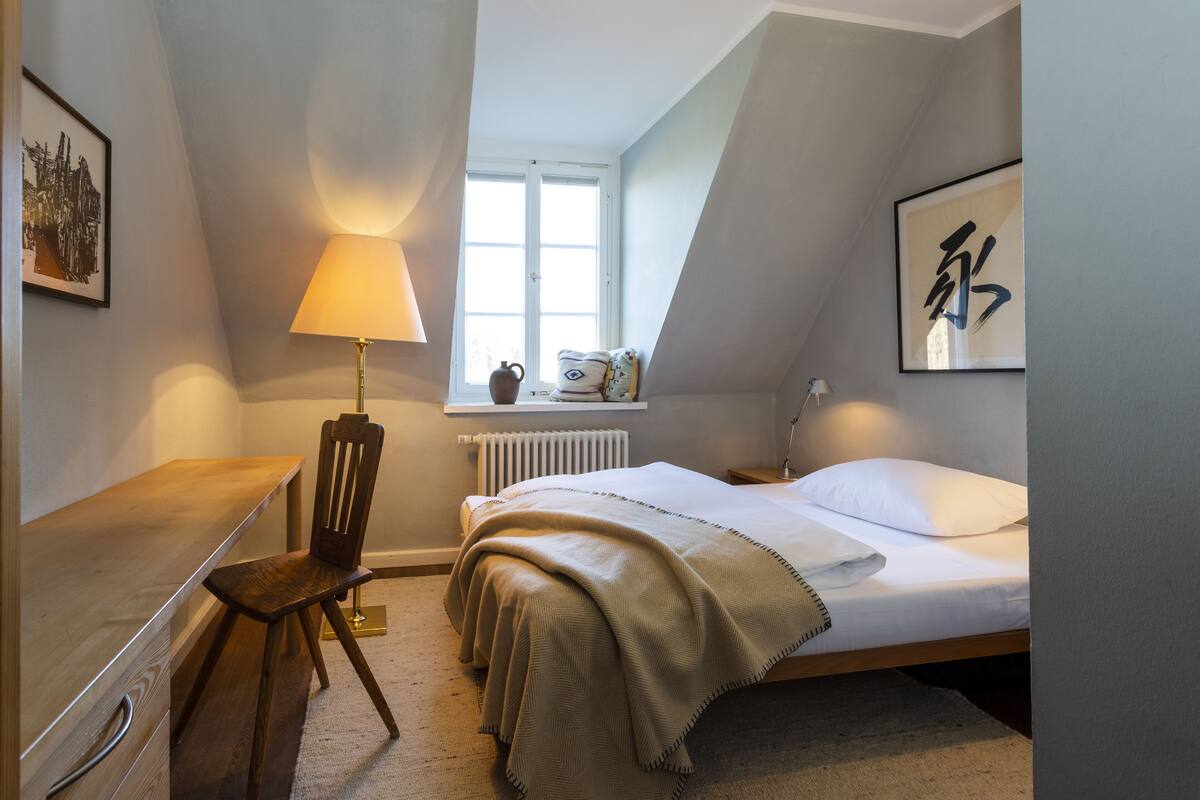 Une chambre à coucher avec un lit, un bureau, une chaise, une lampe et des œuvres d’art accrochées aux murs.