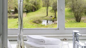 A bathroom with a large window, a white sink, a tall glass vase with a branch, and neatly stacked white towels. - Gut Sonnenhausen (Glonn)