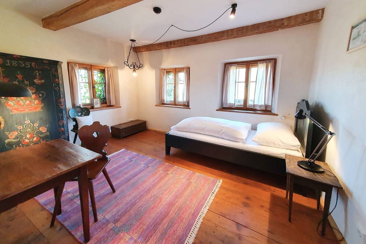 Une pièce confortable avec un lit en bois, un bureau et une chaise. Il y a des fenêtres avec des rideaux et un luminaire au plafond.
