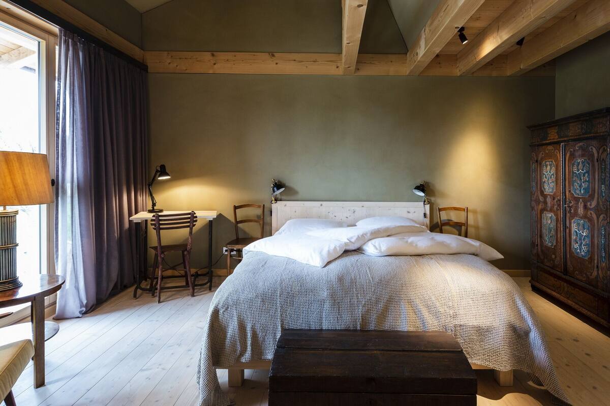 Une chambre à coucher avec une tête de lit en bois, un lit recouvert de draps blancs, une table de chevet en bois, un bureau avec une lampe et une armoire ornée de motifs décoratifs sculptés.