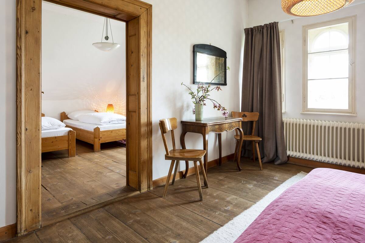 Une pièce avec un plancher en bois, un lit avec une literie blanche, un bureau orné d’un vase de fleurs et une chaise.
