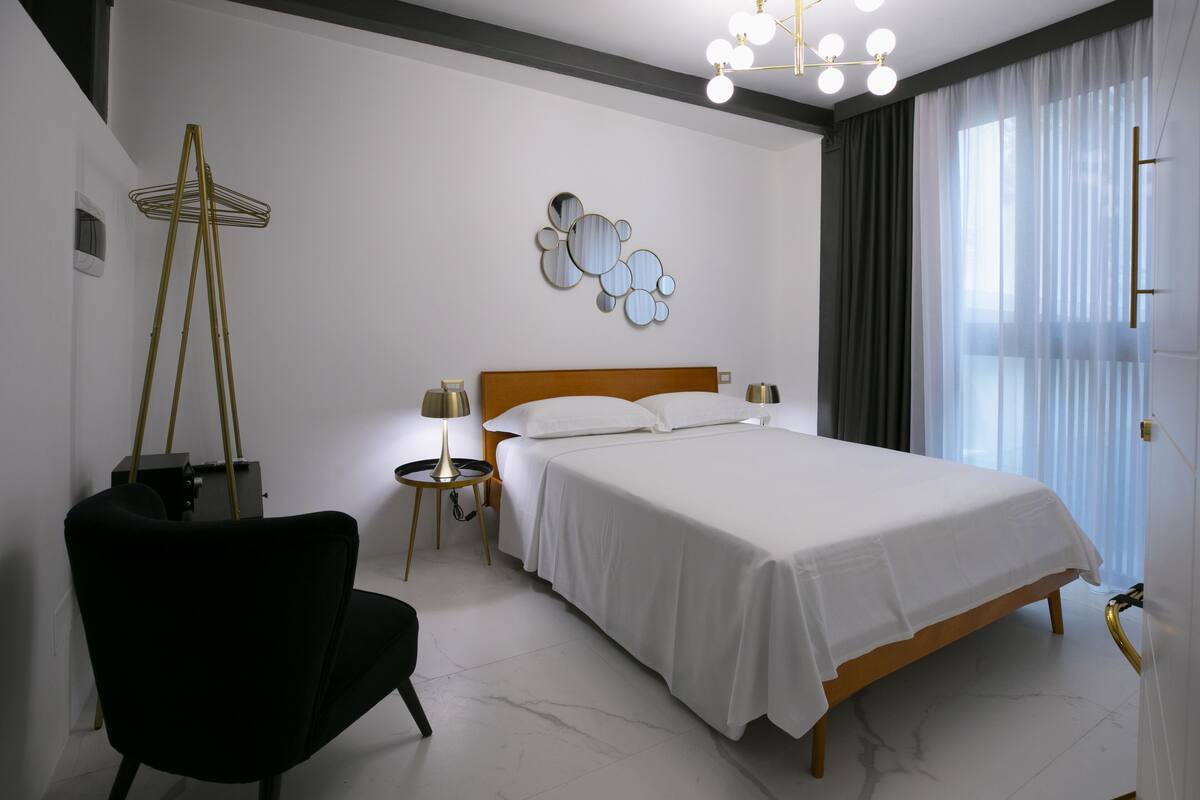 Un dormitorio moderno con una cama de madera, una silla negra, una mesita de noche con lámpara y un elemento decorativo en la pared.
