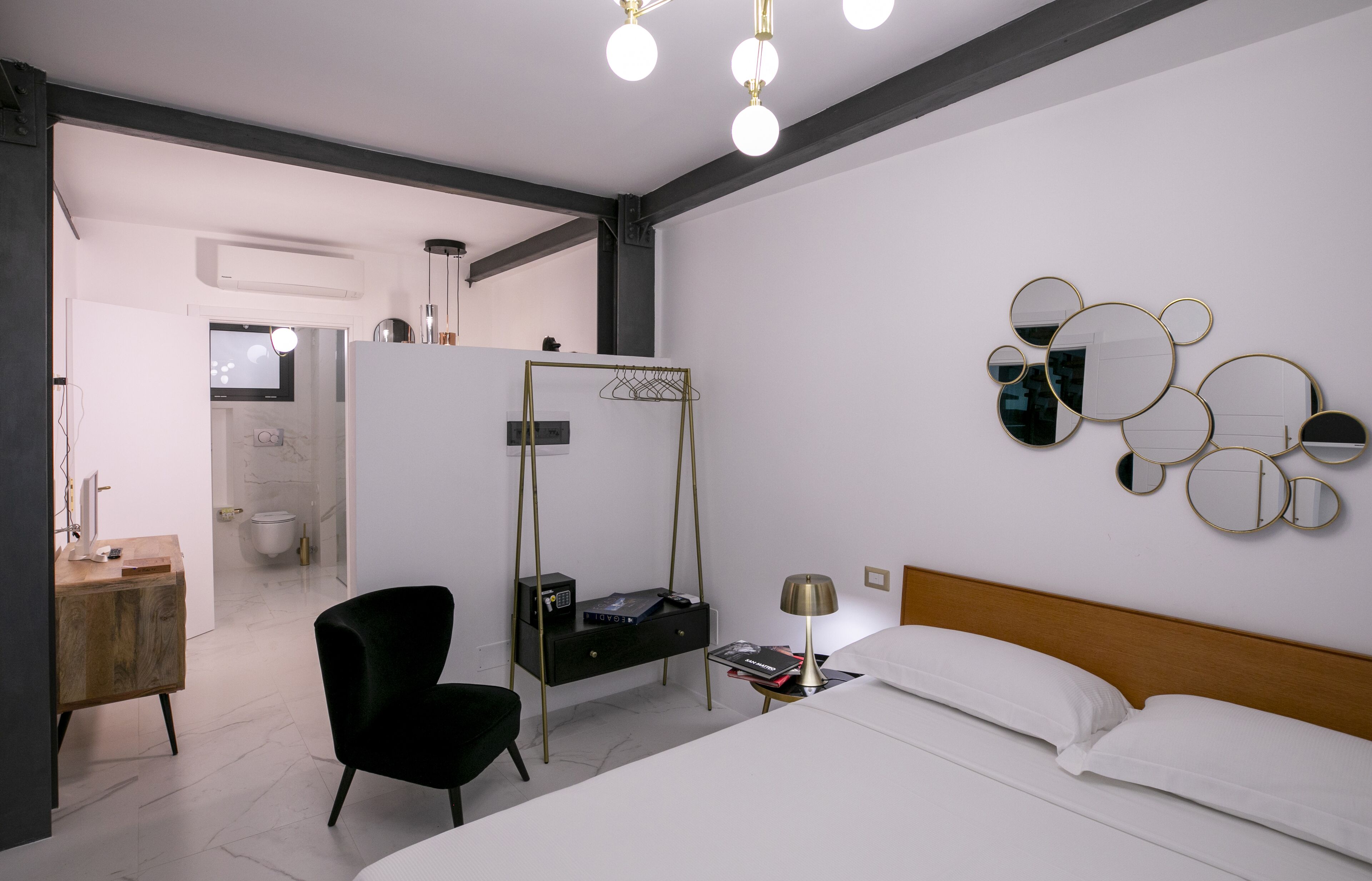 Ein modernes Hotelzimmer mit einem Bett, einem schwarzen Sessel, einem Nachttisch mit Lampe, einem wandmontierten Spiegel und einer kleinen Ablagefläche mit Fernseher und weiteren Gegenständen.