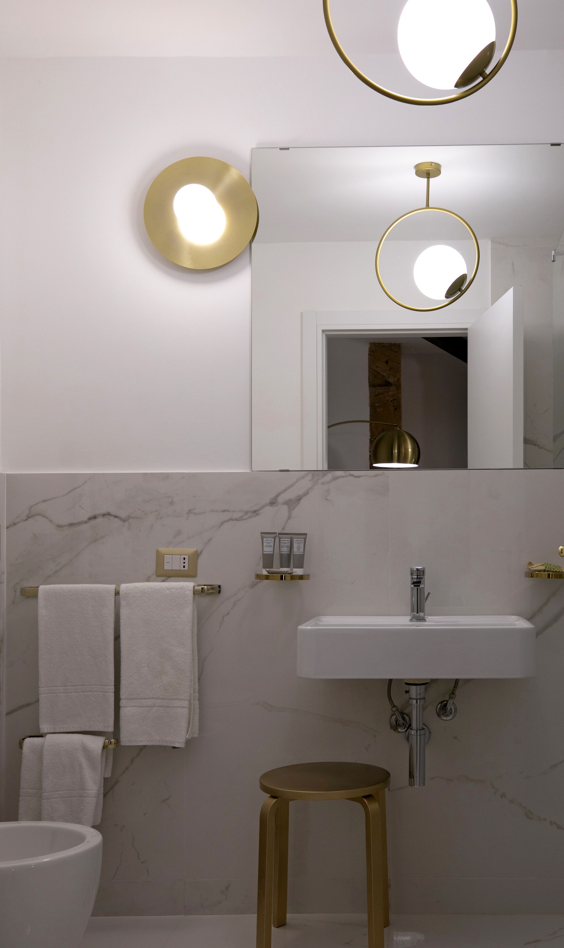Ein modernes Badezimmer mit Waschbecken in Weiß, goldenen Armaturen, Marmorwänden und einem runden Spiegel.