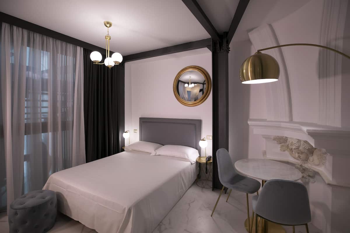 Una habitación de hotel moderna con una cama grande, mesitas de noche con lámparas, un espejo redondo y una pequeña mesa redonda con sillas.