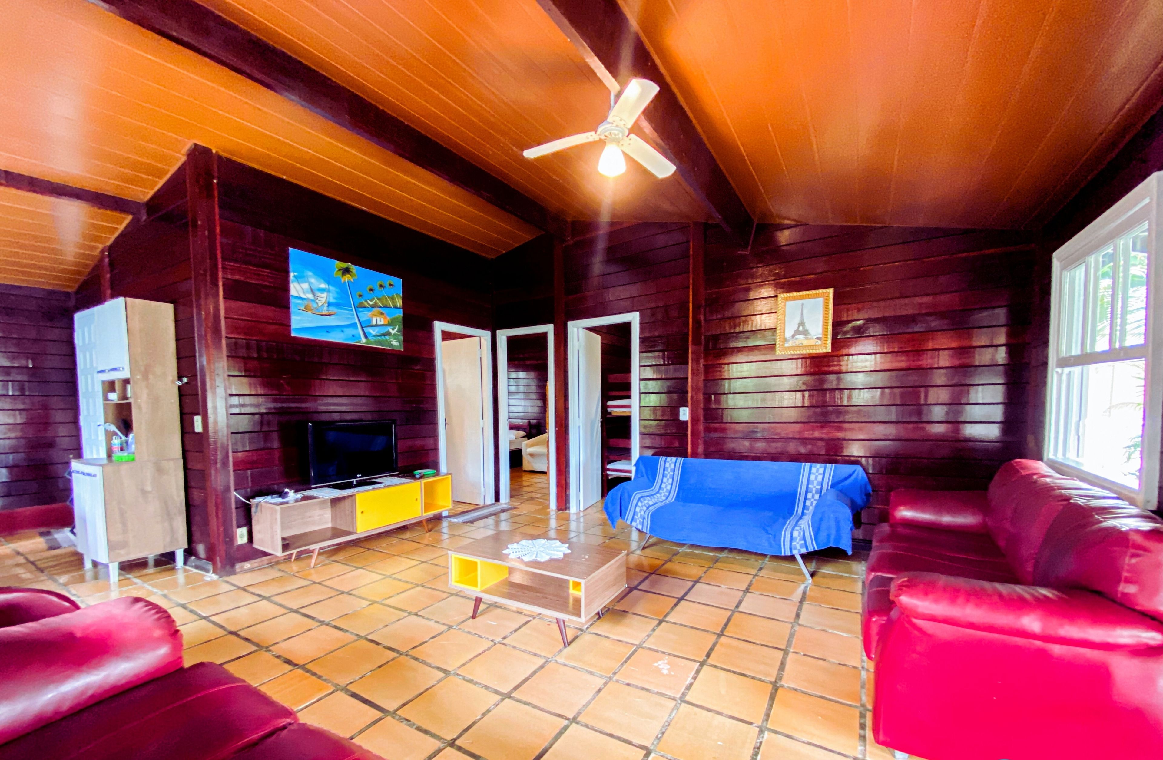 Una sala de estar acogedora con un sofá rojo, un televisor y una mesa de centro. Hay un ventilador de techo y un cuadro en la pared.