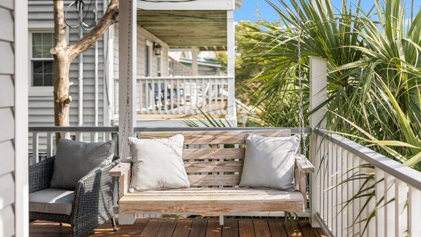 Una terraza de madera con un banco y una silla de mimbre, rodeada por una baranda blanca y plantas de palma.