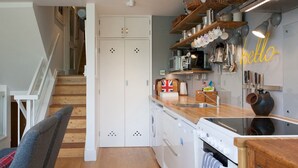 Cocina moderna con armarios de madera, horno integrado y pileta de acero inoxidable. Se ve una escalera que sube a otro nivel, y una repisa con utensilios de cocina y un letrero de neón.