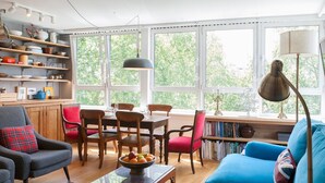 Un salón moderno con un sofá azul, una mesa de comedor de madera y sillas. Hay una estantería llena de libros y una ventana con vistas a árboles.