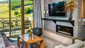 Een moderne woonkamer met een open haard, een flatscreen televisie en uitzicht op een groen landschap door grote ramen.