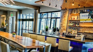 Een bar met een lange houten tafel en verschillende barstoelen, een stenen muur met een hockeyposter, en uitzicht op de stad door grote ramen.