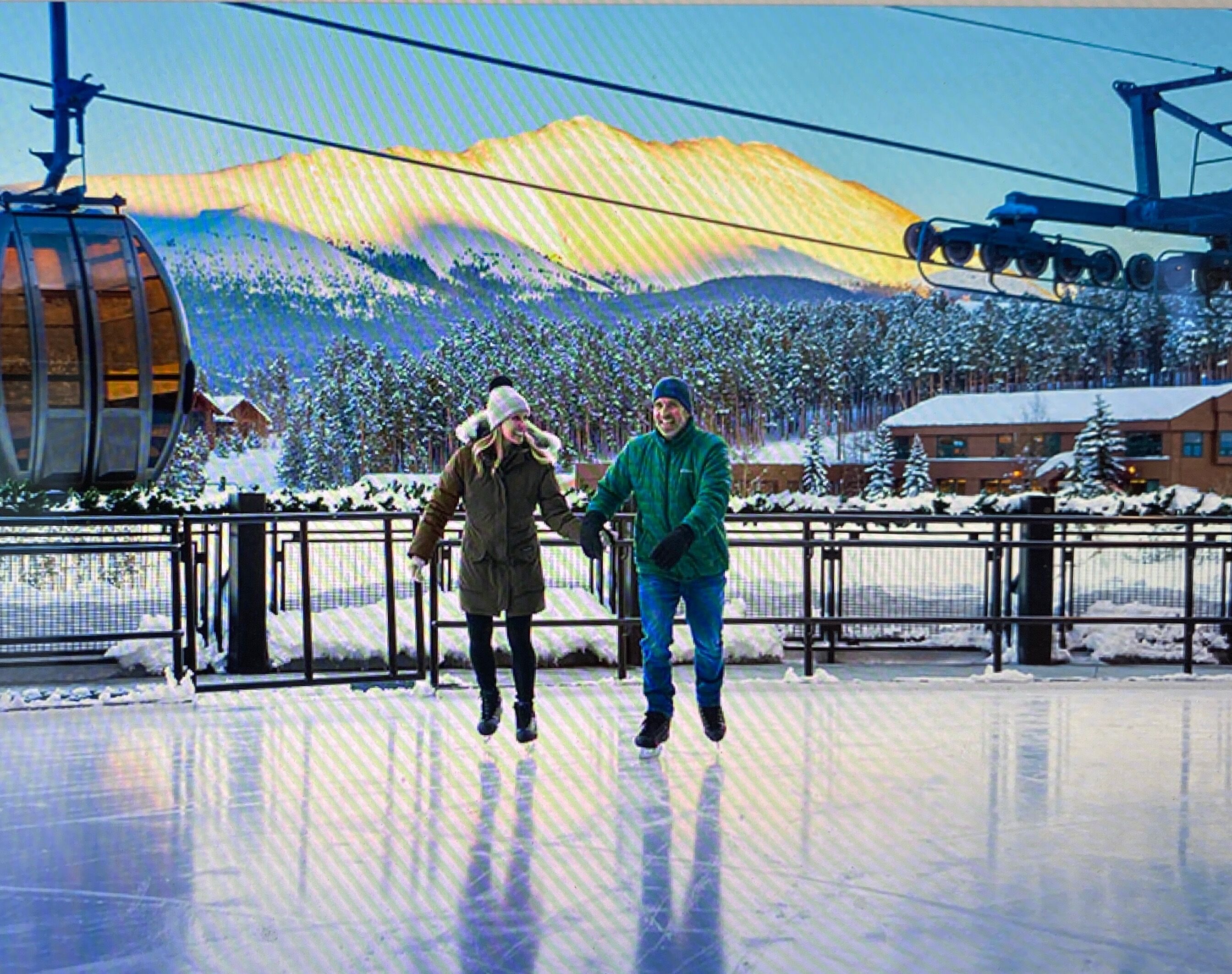 Two people ice skating on a frozen surface with a ski lift and snowy landscape in the background.