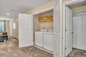 A laundry room with a washing machine and dryer, a drying rack, and a gray couch in a hallway.