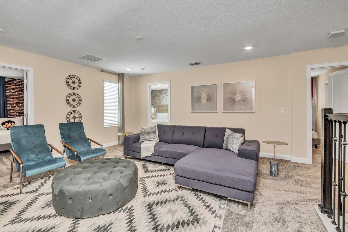 A living room with a sectional sofa, two armchairs, a round ottoman, and decorative wall clocks.