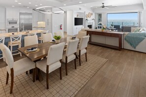 A modern kitchen with white cabinetry, a dining area with a wooden table and chairs, and a view of the ocean through a large window.