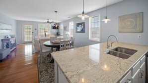 A modern kitchen with a large island, pendant lighting, and a dining area with a table and chairs. - Classic Beach Cottage on Oceanside of Wrightsville Beach-Upper Unit (Wrightsville Beach)