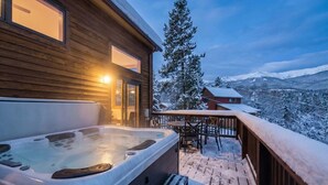 Une cabane en bois dotée d’un bain à remous sur une terrasse, entourée d’arbres et de montagnes couverts de neige.