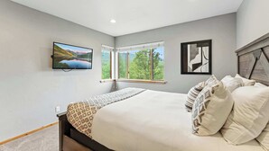 Une chambre à coucher avec un lit, un téléviseur fixé au mur, une fenêtre donnant sur des arbres et un tableau encadré accroché au mur.