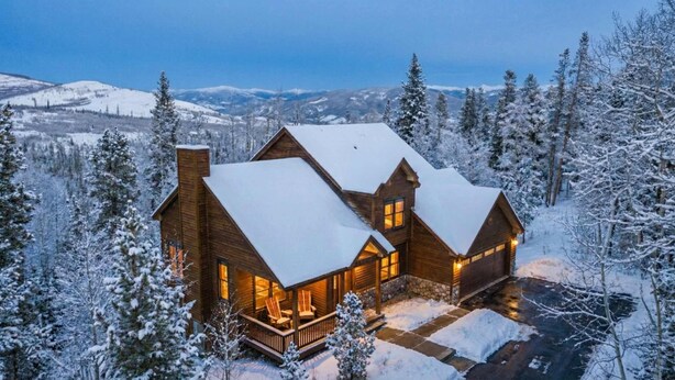 Une cabane en bois dont le toit est recouvert de neige, entourée d’arbres chargés de neige et avec des montagnes en arrière-plan.