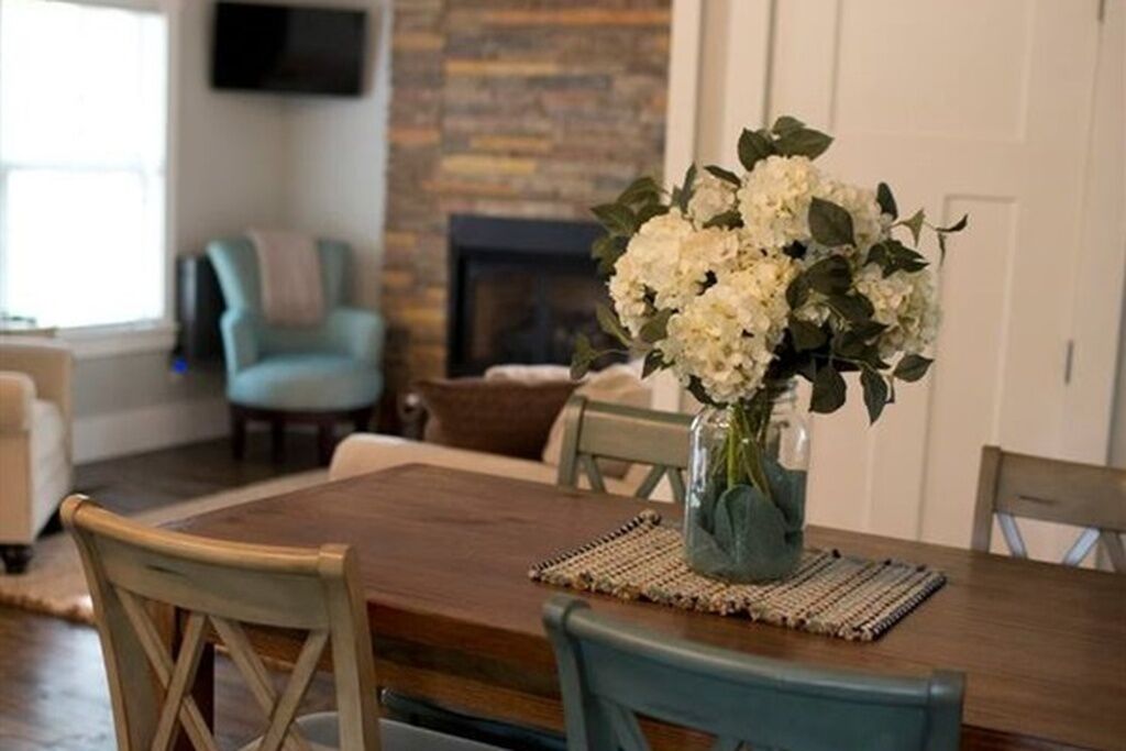 A dining table with chairs, a vase with flowers, and a fireplace in the background.