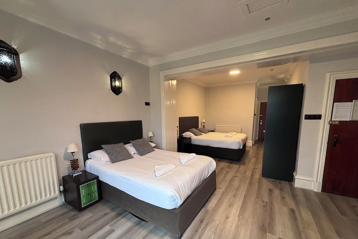 A hotel room with two beds, a radiator, a nightstand with a lamp, and a wall-mounted light fixture.