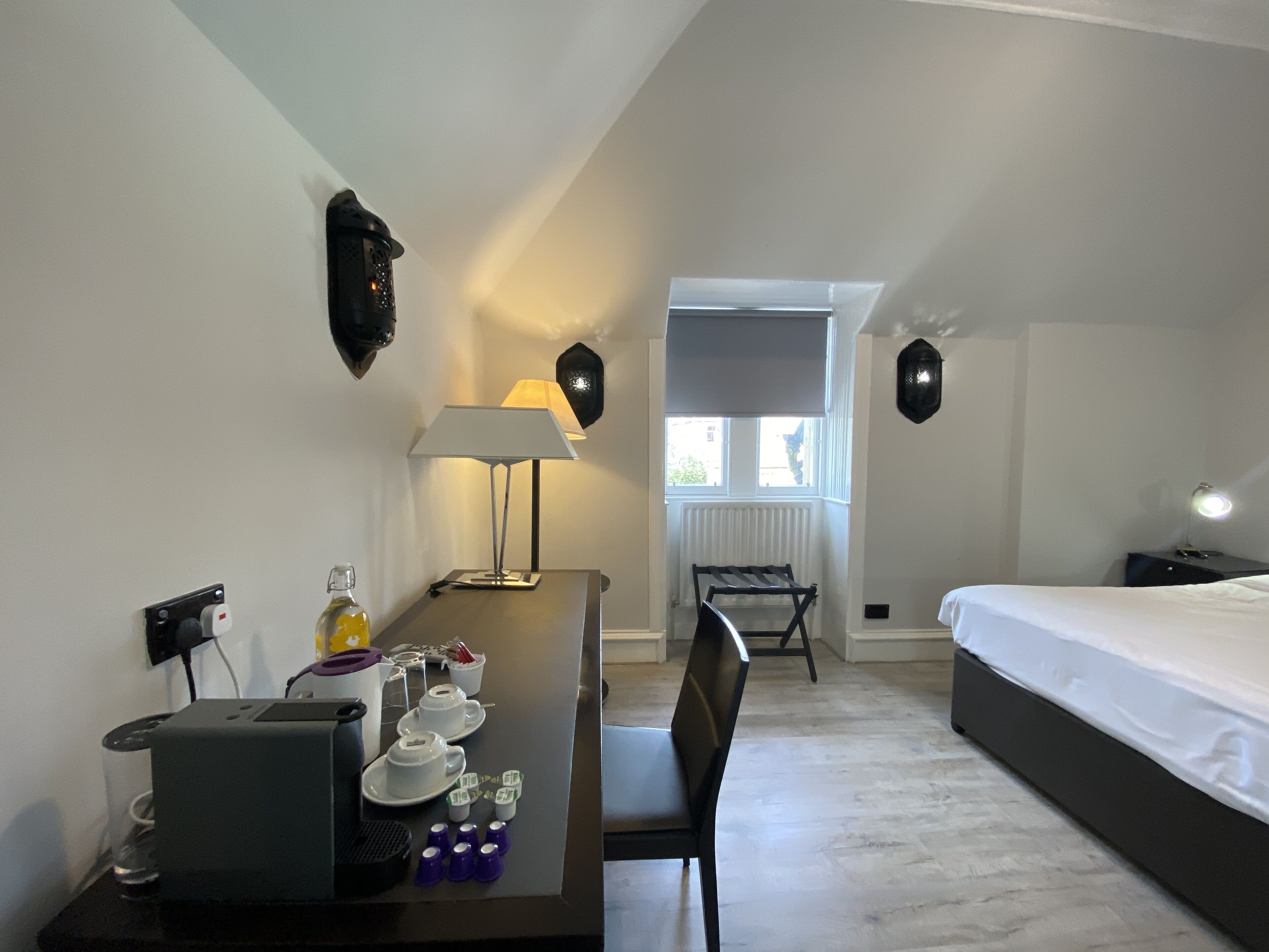 Habitació d'hotel amb un llit, un escriptori amb una làmpada, una cadira, un radiador i una finestra.