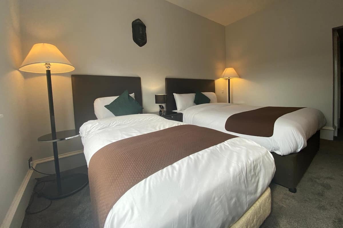 A hotel room with two beds, each with a brown throw blanket, white bedding, and a nightstand with a lamp.
