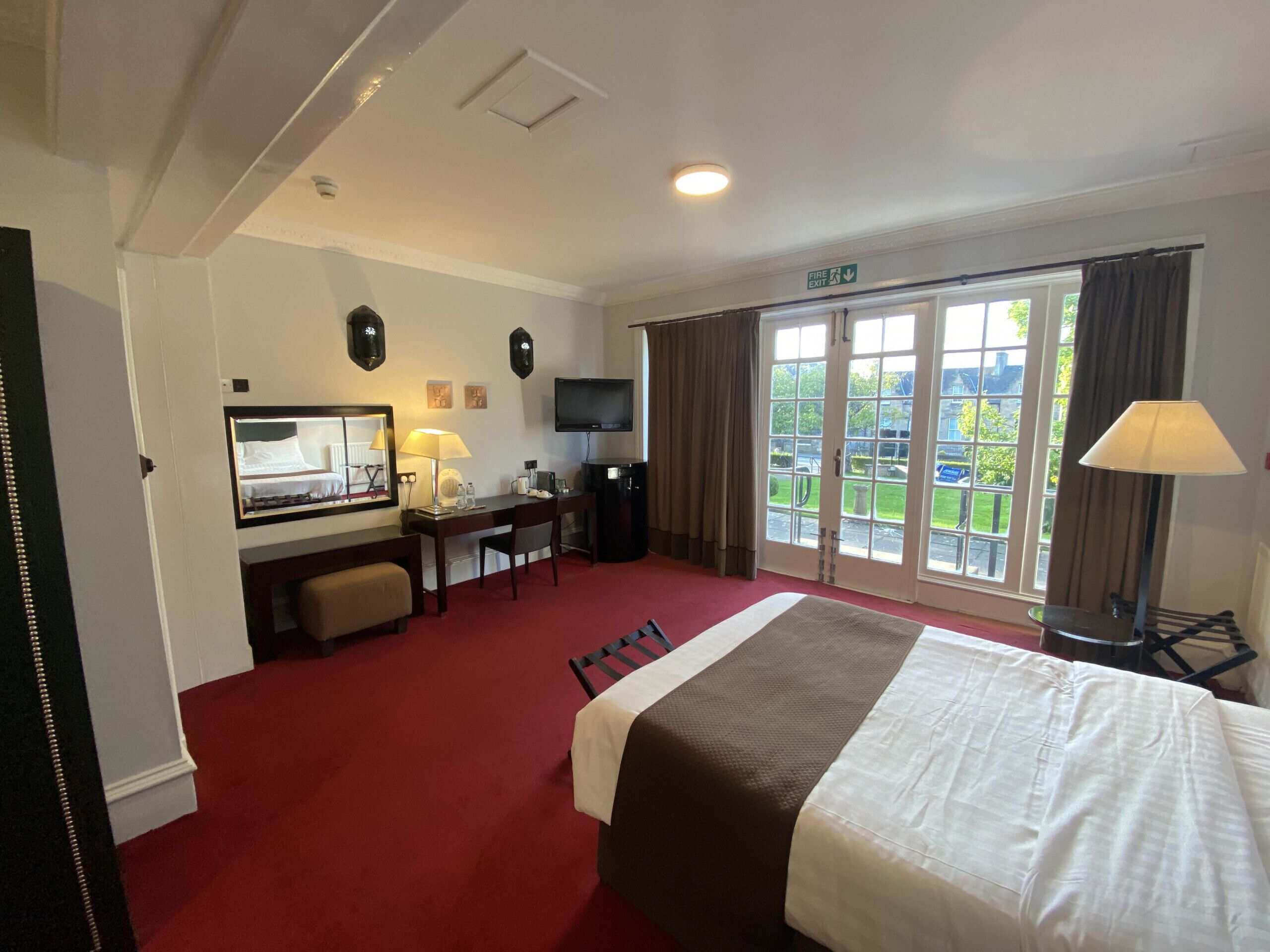 A hotel room with a bed, desk, chair, television, and a view of the outdoors through large windows.