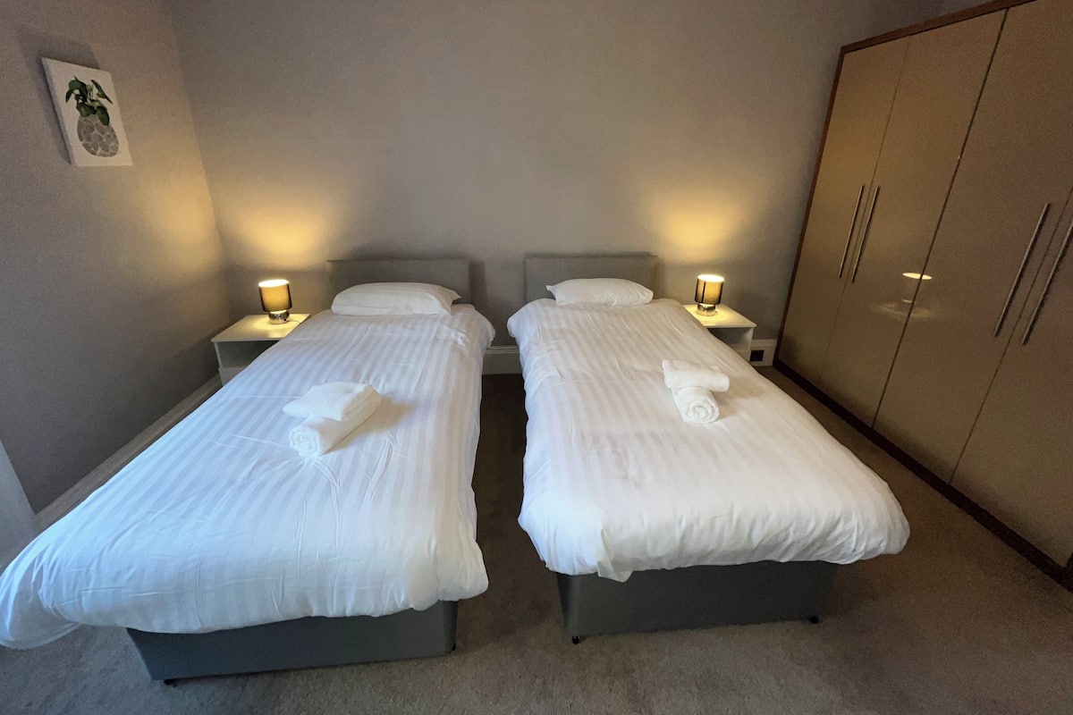A hotel room with two single beds, each with a bedside table and lamp, a wardrobe, and a wall-mounted picture.