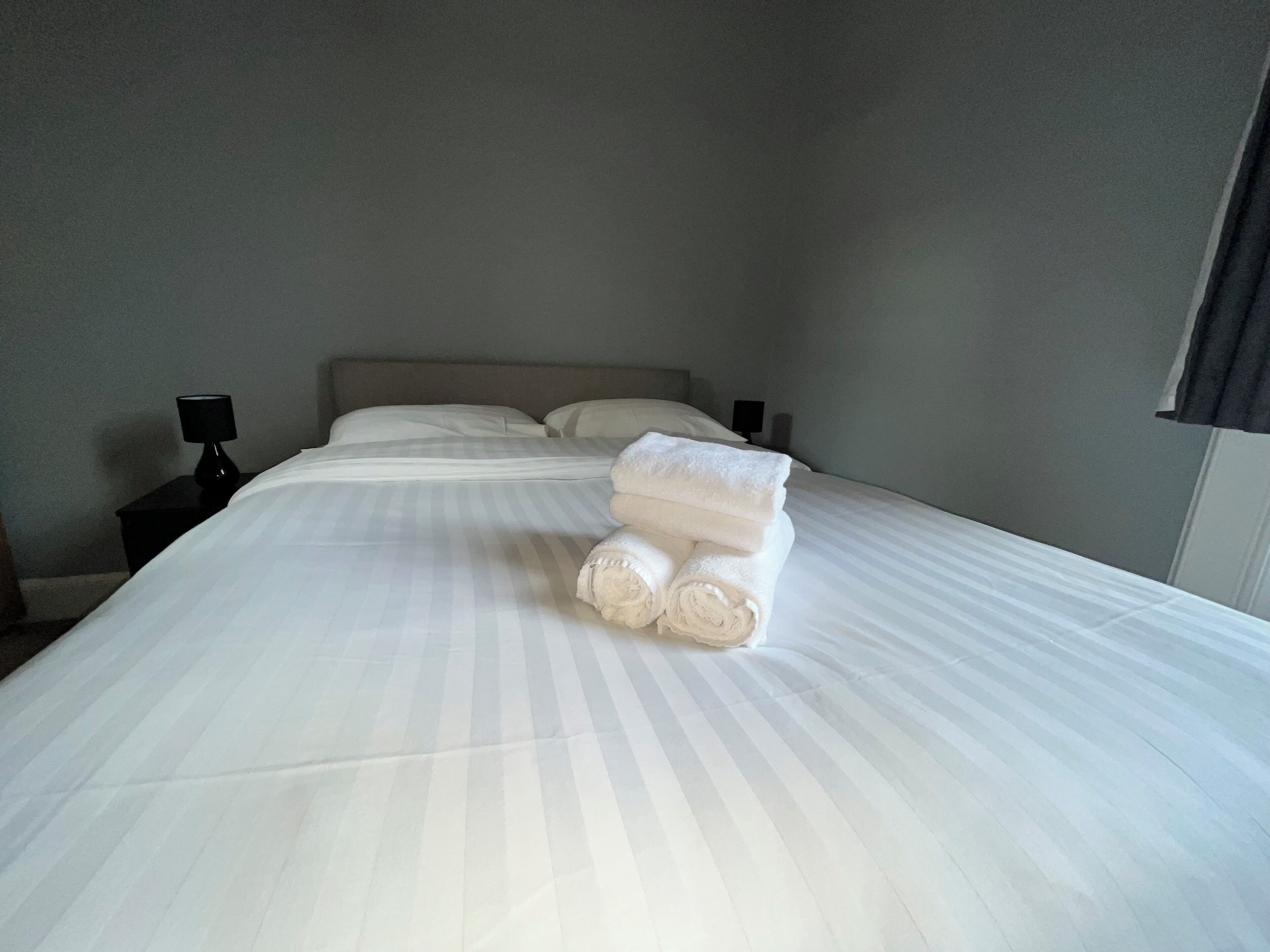 A neatly made bed with white linens and rolled towels, a bedside lamp, and a dark nightstand.