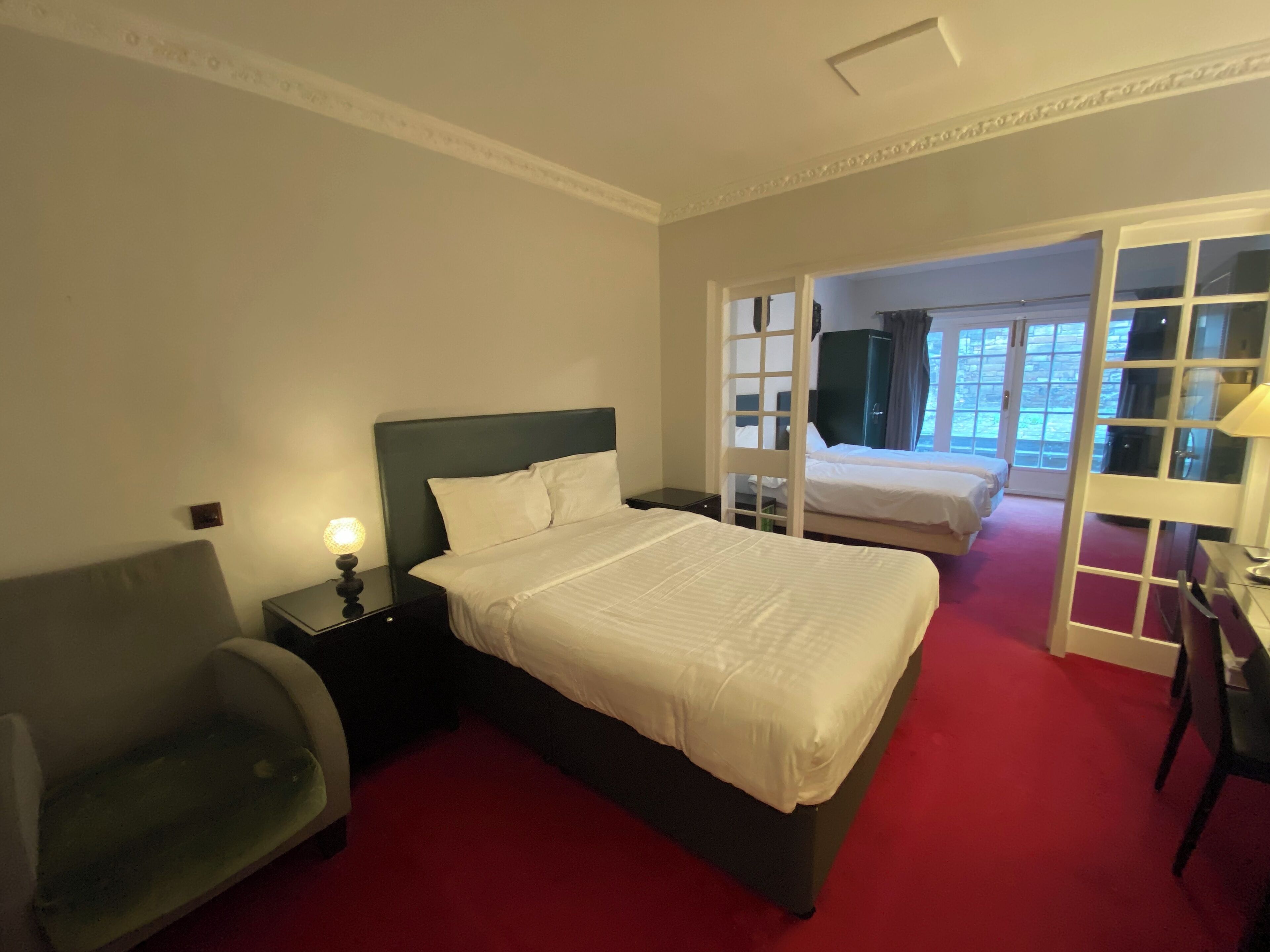 A hotel room with a large bed, a chair, a desk, and a view through a glass door to a balcony.