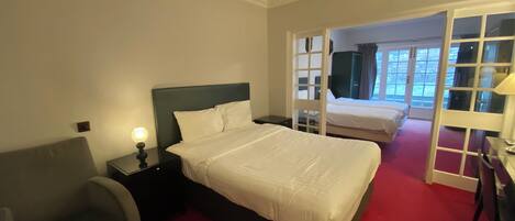 A hotel room with a large bed, a chair, a desk, and a view through a glass door to a balcony.