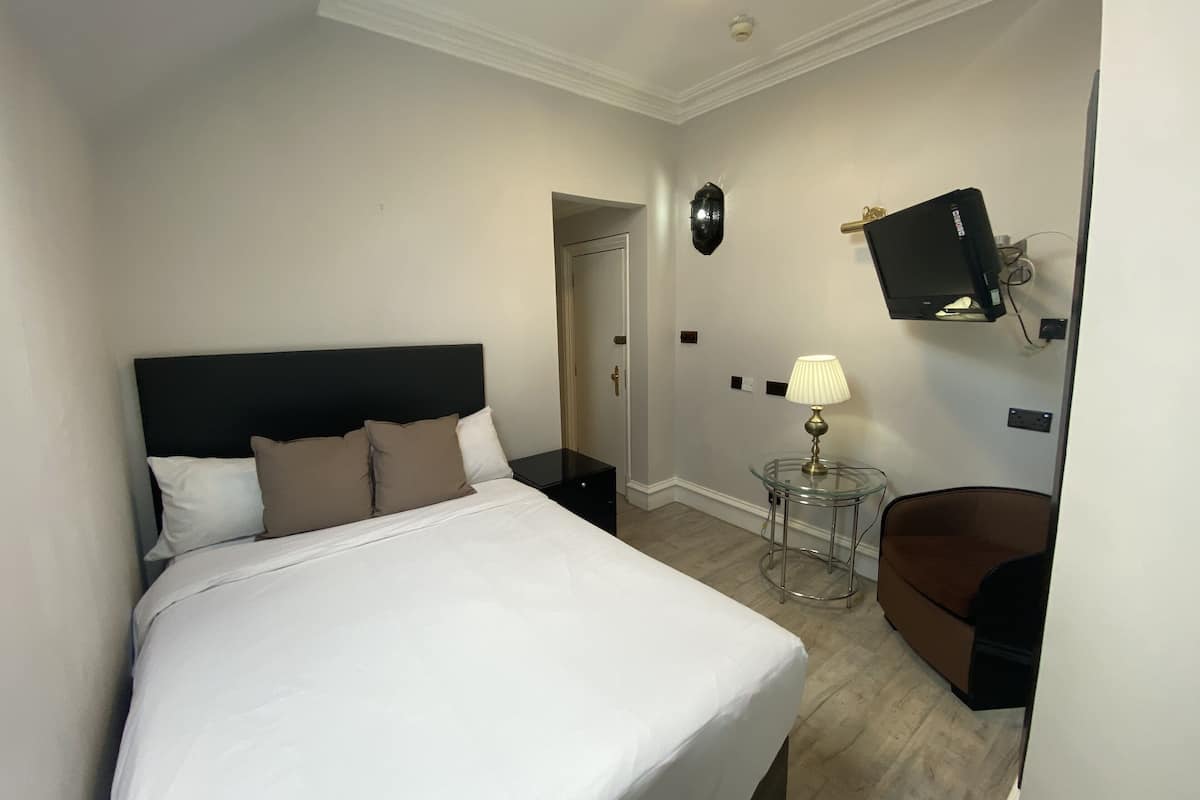 A hotel room with a bed, a desk, a chair, a TV mounted on the wall, and a lamp on a side table.