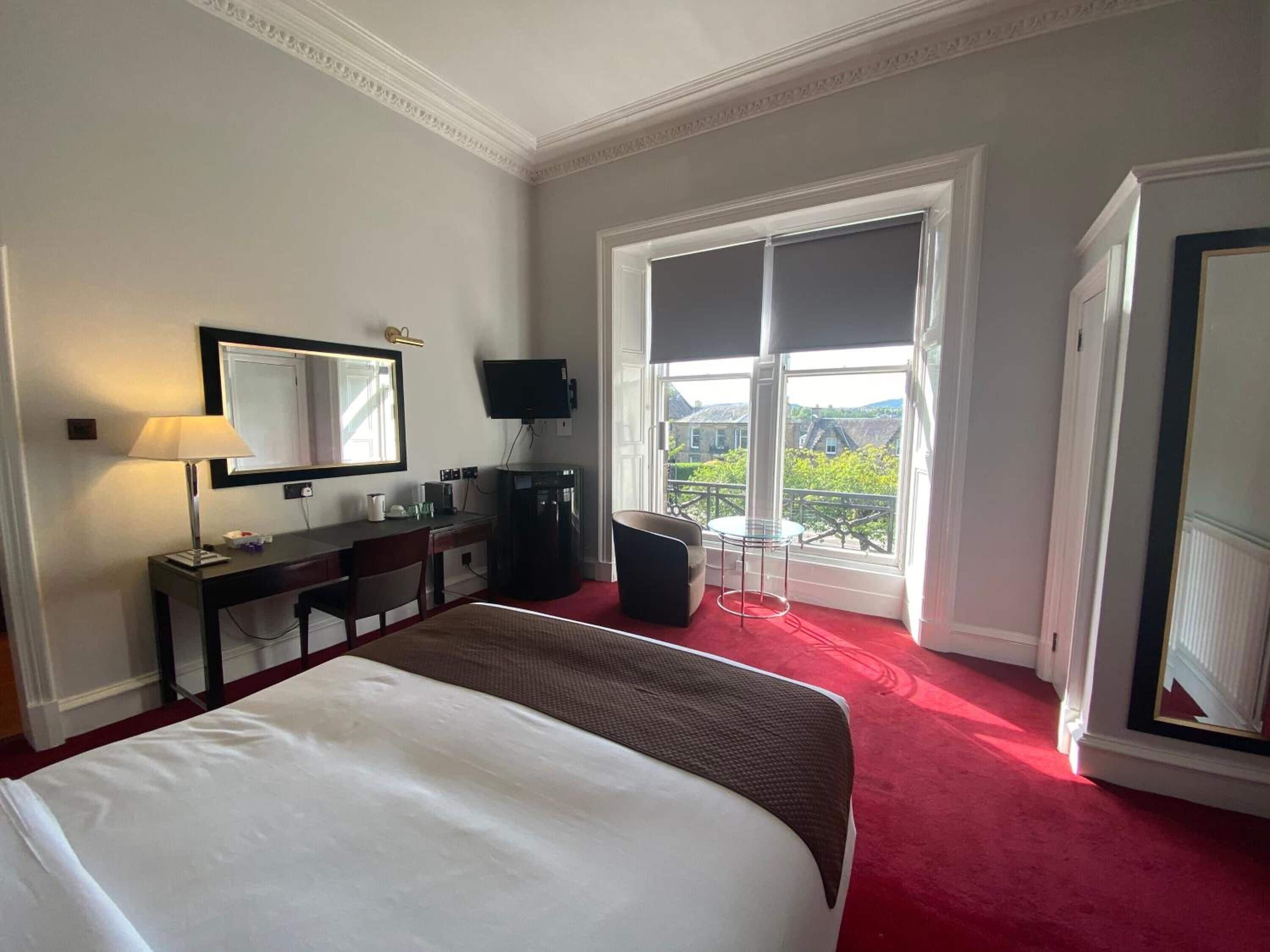Ein Hotelzimmer mit Bett, Schreibtisch mit Stuhl, Fernseher, Kaffeemaschine und einem großen Fenster mit Außenblick.