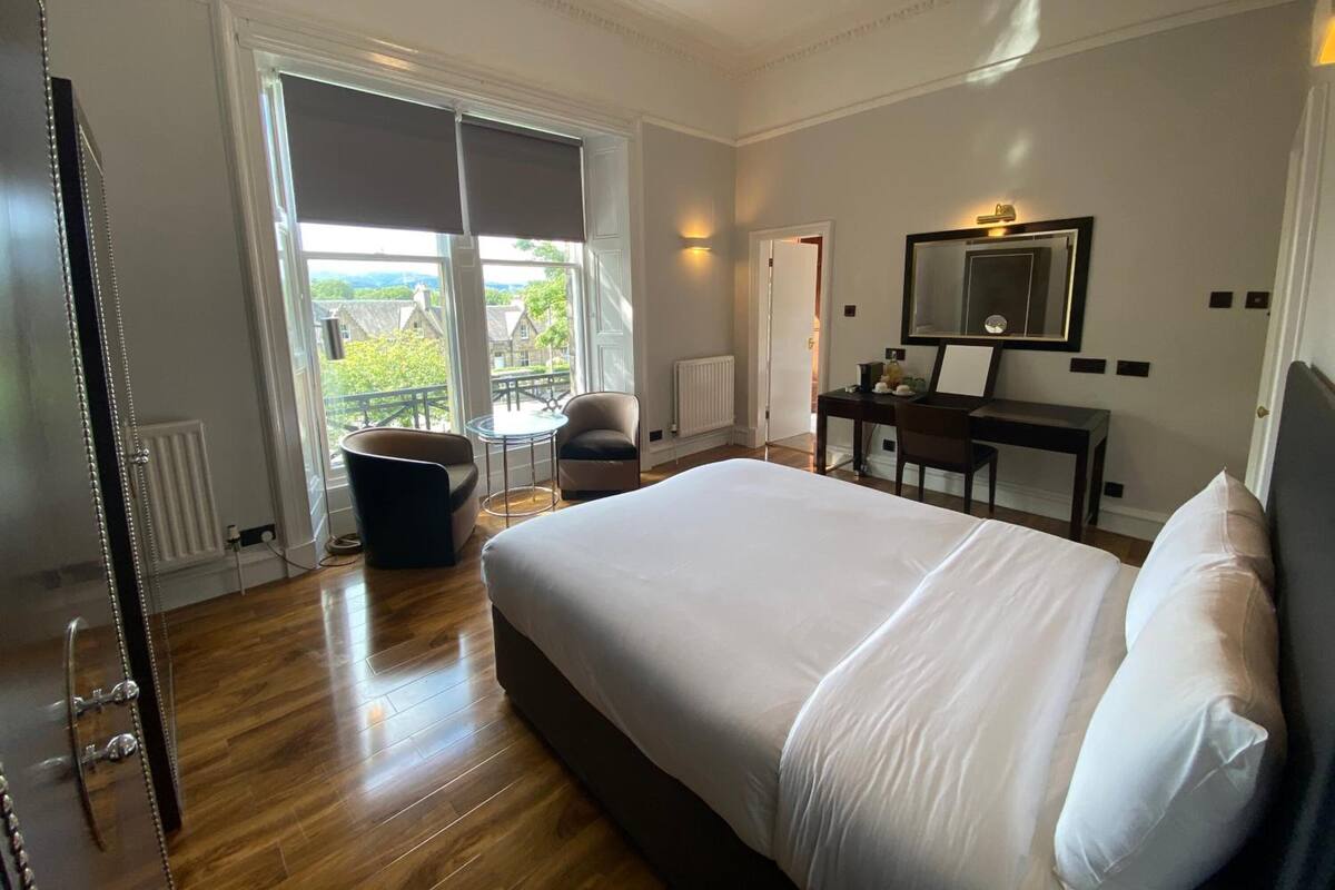 A hotel room with a large bed, a desk, a chair, a small table, and a view of the outdoors.