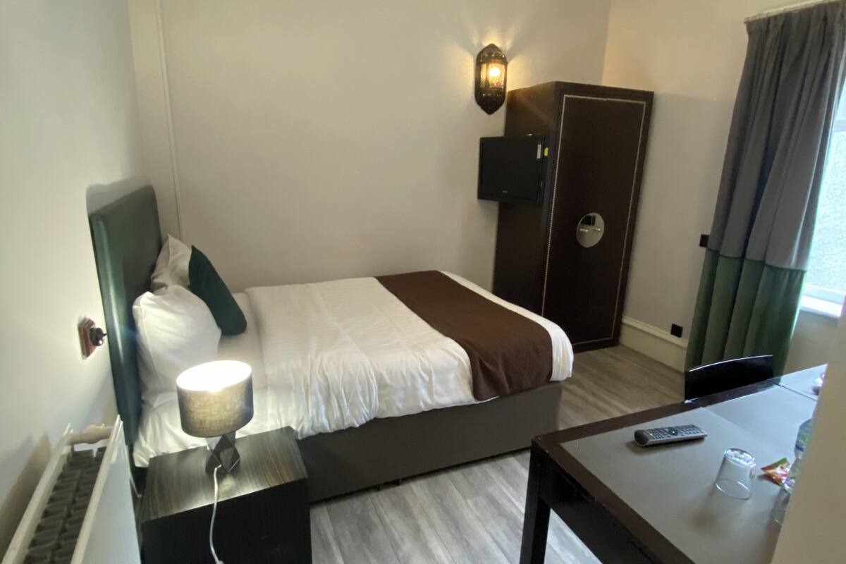 A hotel room with a bed, bedside table with a lamp, a desk with a remote control, and a door with a lock.