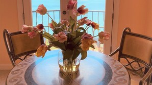 A round table with a glass top and a vase of flowers, positioned in front of a door leading to a balcony with a view of the sea. - Hidden apartment at Villa del Sol ~ Amazing Sunsets -Spacious Luxury (Austin)