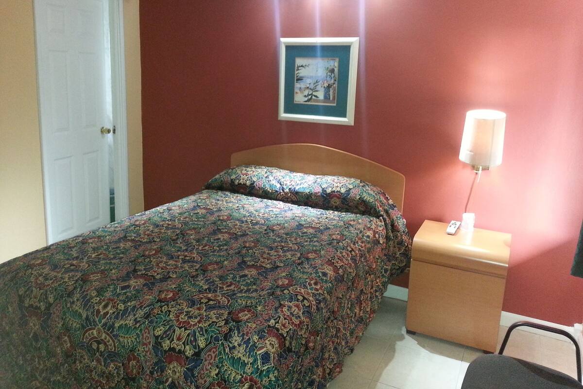 Un dormitorio con cama, mesita de noche, lámpara, un cuadro enmarcado en la pared y una puerta.