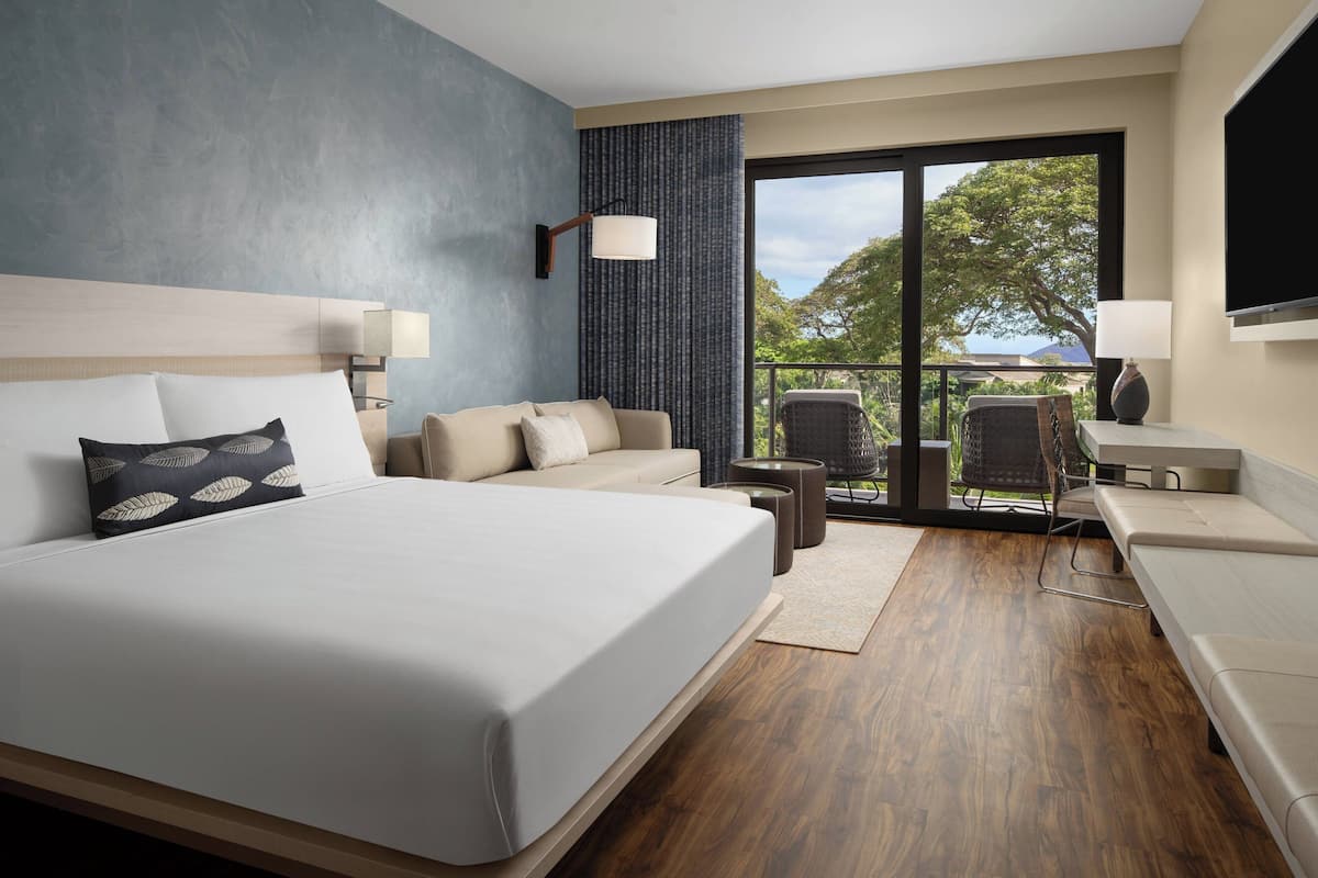 Una habitación de hotel moderna con una cama grande, un sofá, un escritorio y una silla. Cuenta con un amplio ventanal con vistas a árboles y una montaña.