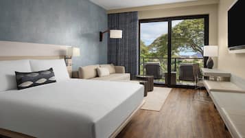 Una habitación de hotel moderna con una cama grande, un sofá, un escritorio y una silla. Cuenta con un amplio ventanal con vistas a árboles y una montaña.