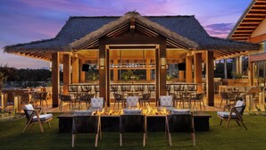 Un espacio lounge al aire libre con techo de paja, fogón y varios conjuntos de asientos.