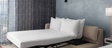 A modern hotel room with a single bed, a bedside table, a lamp, and a view of the outside through curtains.