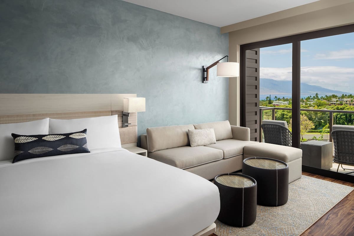Habitación de hotel con una cama grande, un sofá, una mesita y vistas a un balcón con paisaje exterior.