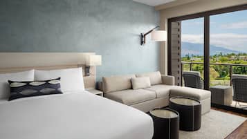 A hotel room with a large bed, a sofa, a small table, and a view of a balcony with outdoor scenery.